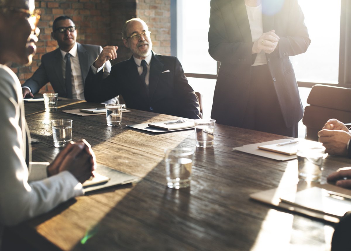 men in suits sitting at a meeting table with a legal pad and glass of water in front of each attendee