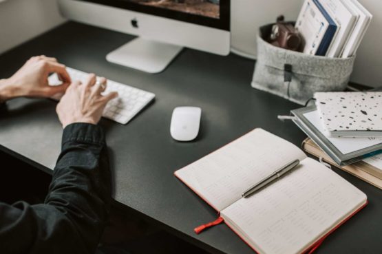 person working on a Mac with an open diary and pen beyond the mouse