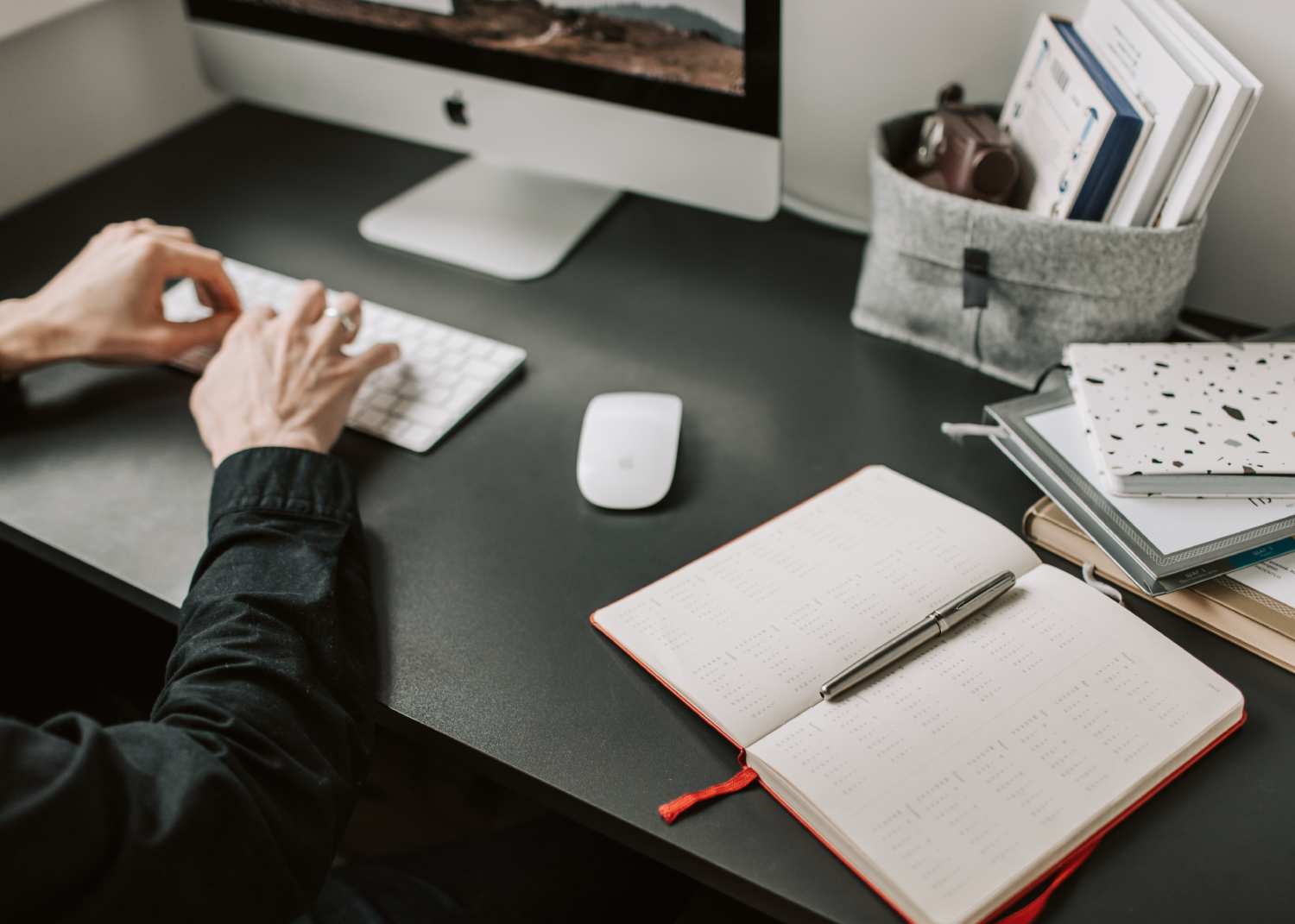 person working on a Mac with an open diary and pen beyond the mouse